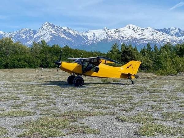 A yellow Maule aircraft is parked in a field with mountains in the background.