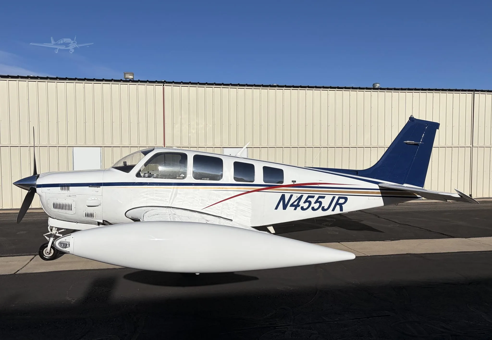 A white single-engine propeller aircraft with blue and red stripes, registration N455JR, parked on an tarmac in front of a hangar.