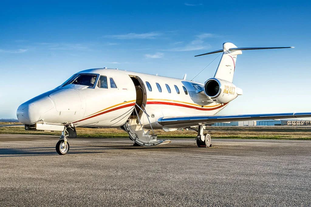 A white business jet with red and gold stripes (N24KT) is parked on a tarmac with its main cabin door open and stairs extended.