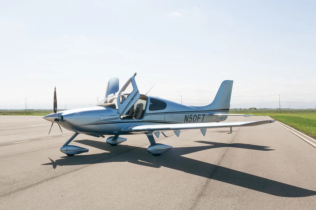 A side view of the light blue Cirrus SR22 aircraft with its gull-wing doors open, parked on an airfield.