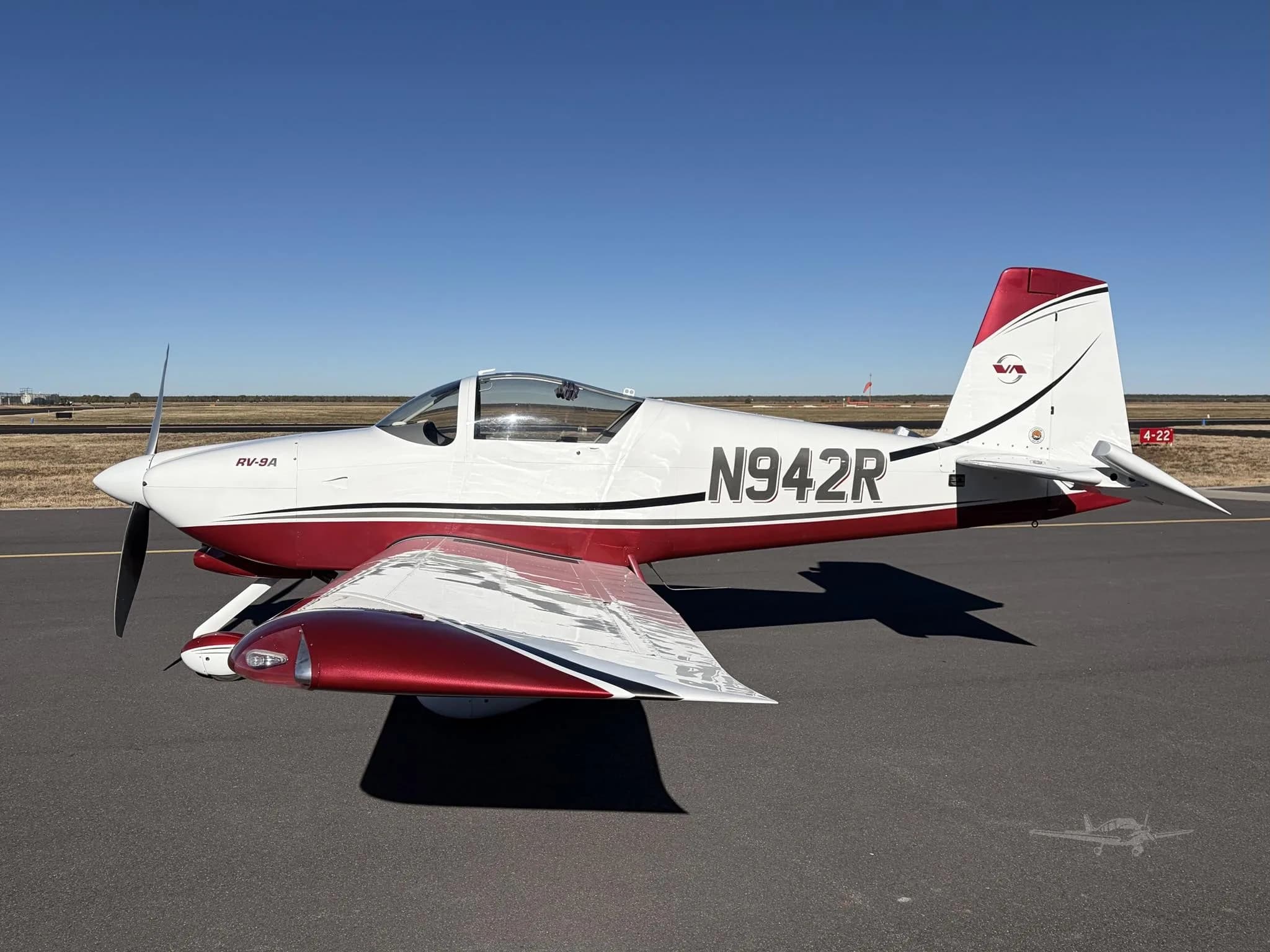 A white and red Van's RV-9A aircraft with tail number N942R is parked on an airport tarmac under a clear blue sky.