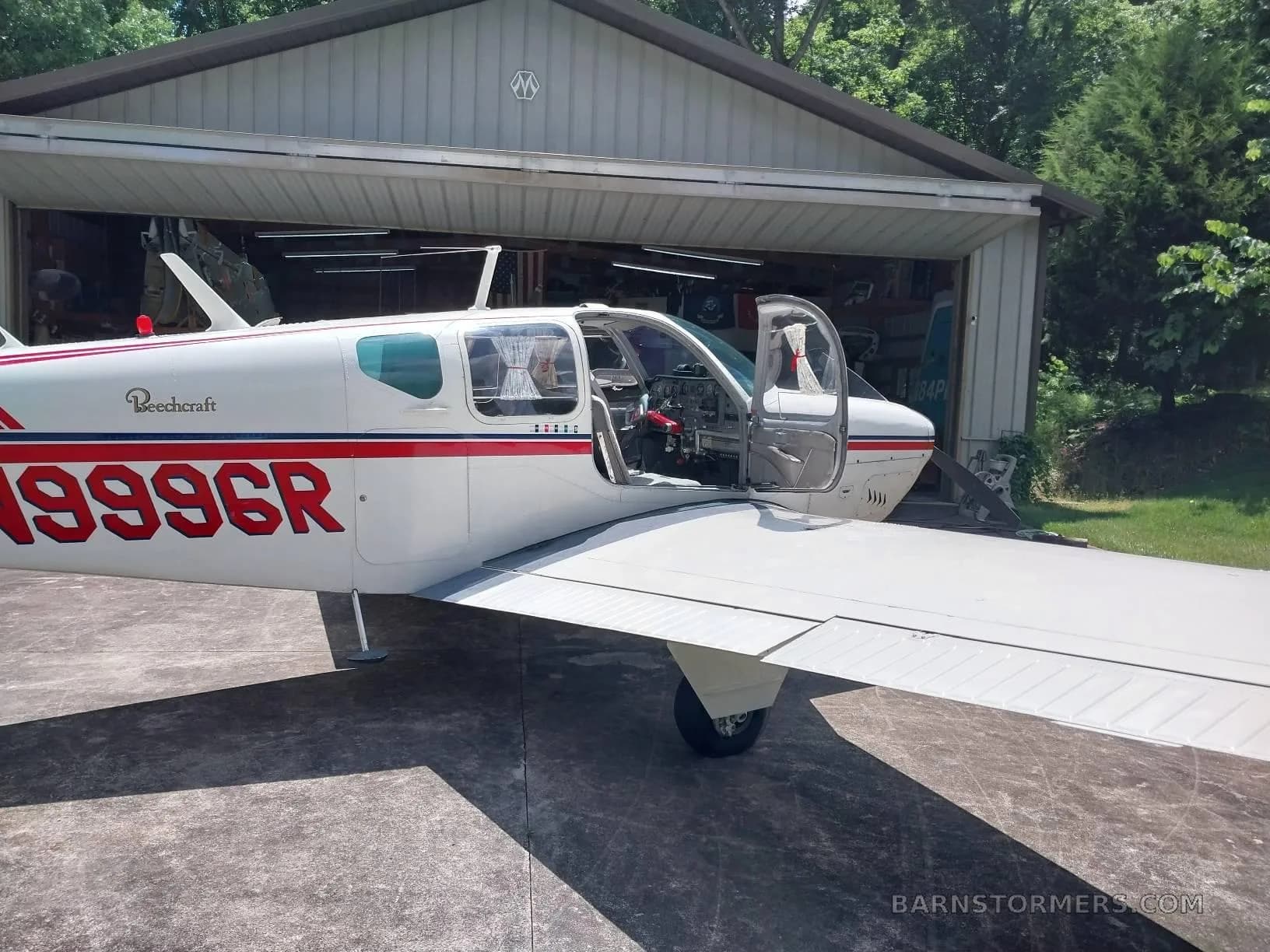 A white Beechcraft Bonanza with red and blue stripes, registration N999CR, parked with its door open next to a hangar.