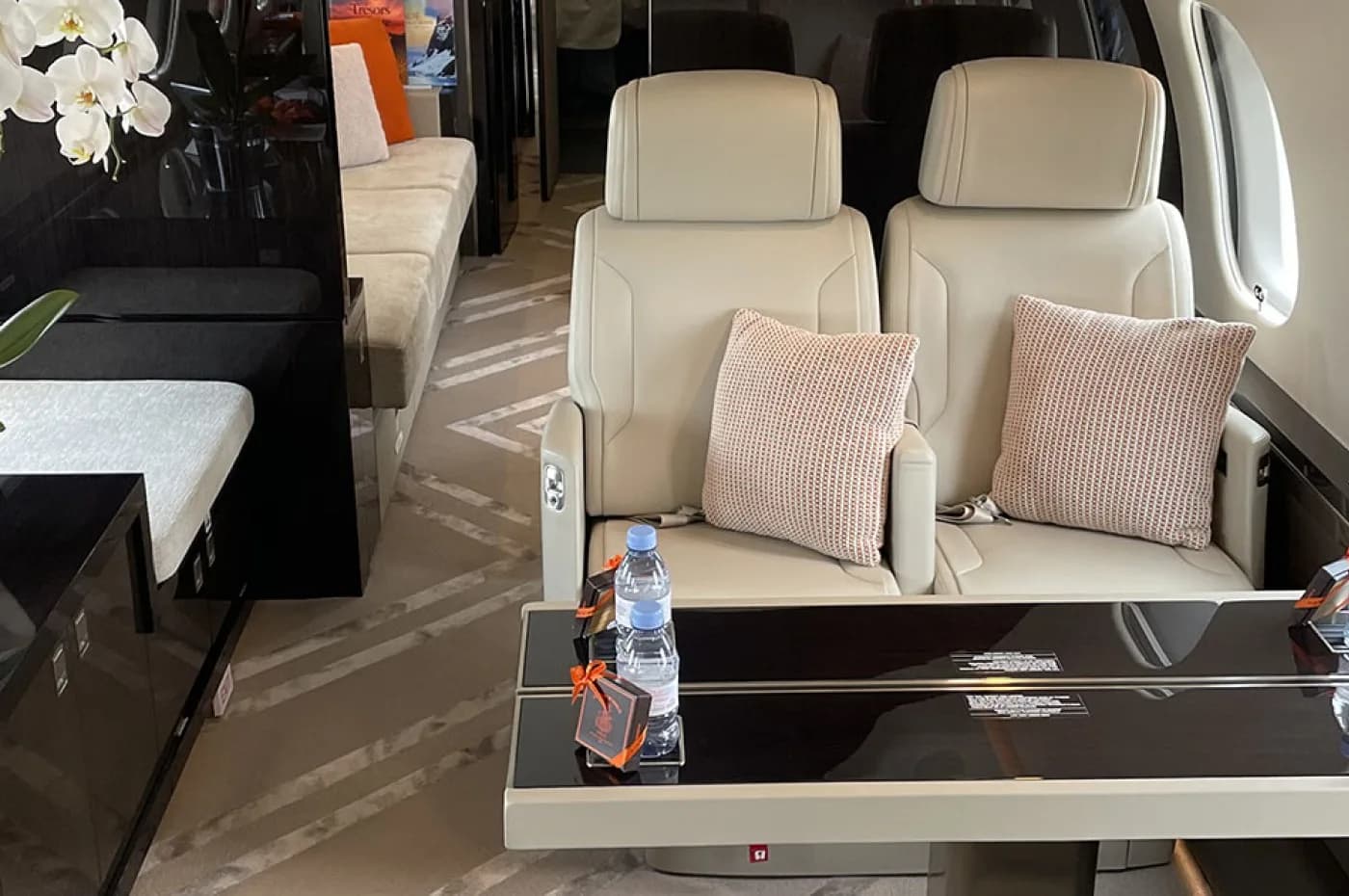 A section of a private jet's cabin interior with two light-colored leather seats, a dark table with water bottles, and a sofa in the background.