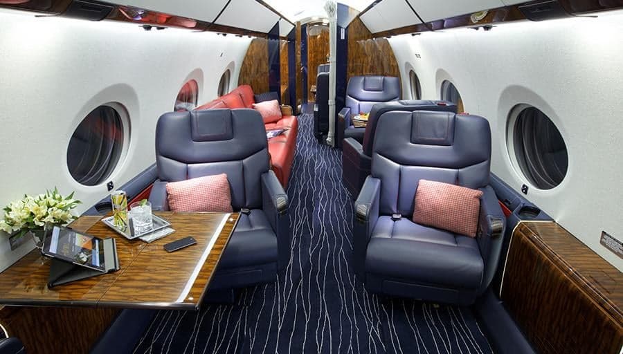 A wide view of the private jet's cabin interior, showing blue leather seats, a dining table, and a red sofa in the background.