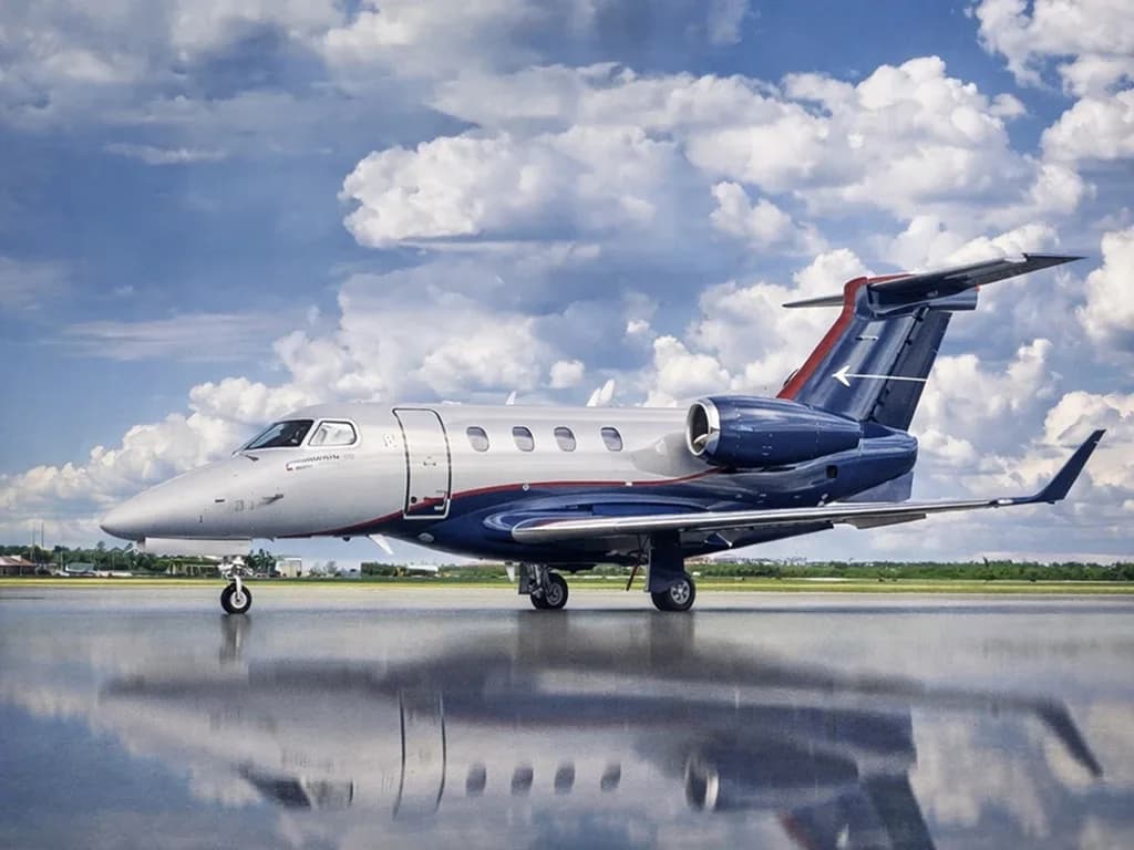 A silver and dark blue Embraer Phenom 300 business jet with a red tail accent sits on a wet tarmac under a cloudy sky.