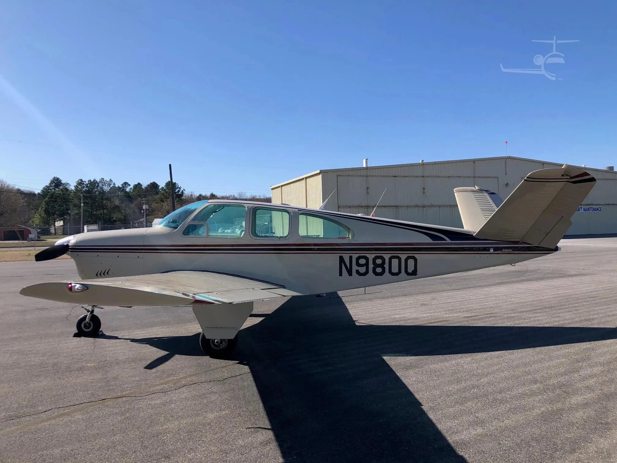 A side view of a beige Beechcraft Bonanza N980Q with red and black stripes parked on an asphalt apron in front of a hangar.