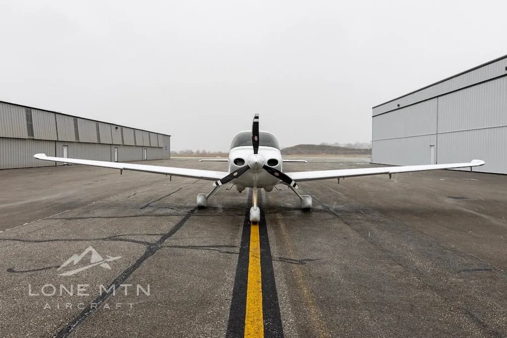 A front view of a white Cirrus SR22 aircraft, tail number N57PX, parked on an airport tarmac between two hangars.