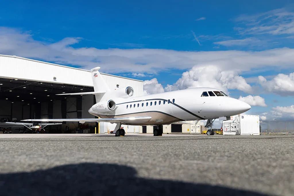A white Dassault Falcon business jet with a brown stripe, registration C-FMCL, parked in front of a hangar.