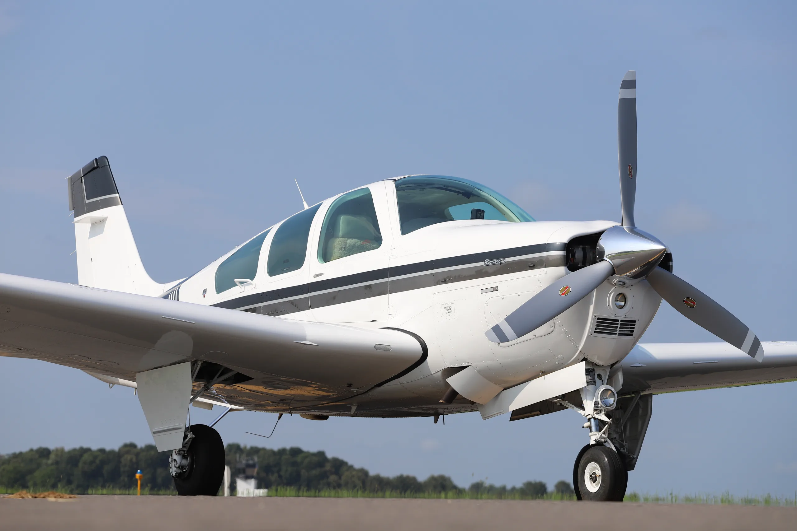 A white Beechcraft Bonanza with a dark grey stripe, seen from a low front-side angle on an airfield.