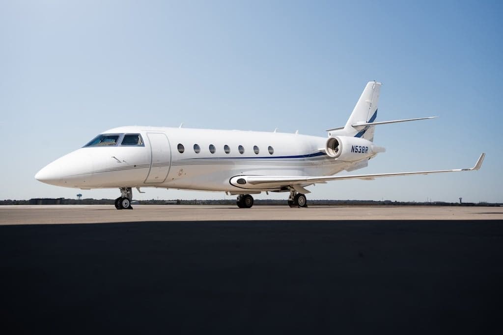 A white business jet with a blue stripe, tail number N538R, parked on an airfield under a clear sky.