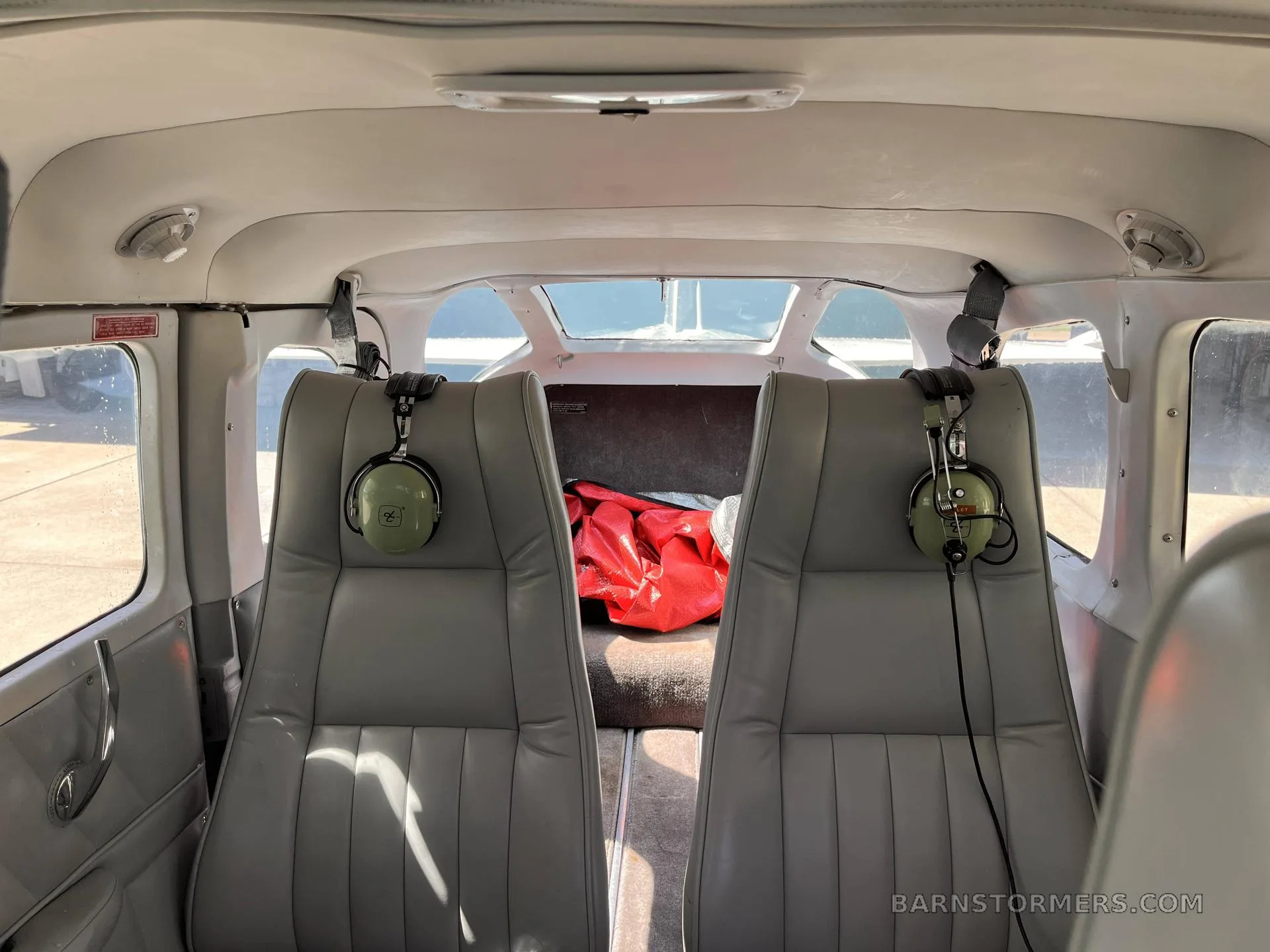 The rear cabin of an aircraft with two light gray seats, each with a green headset, and a red bag in the baggage area.