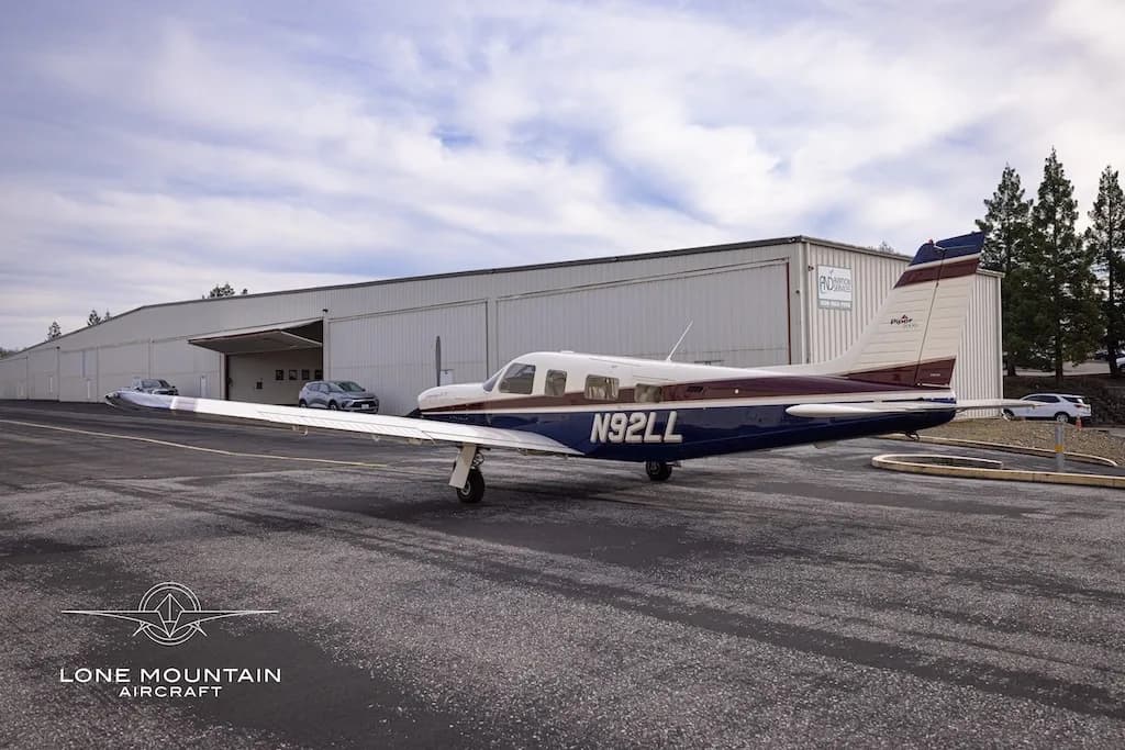 A three-quarter rear view of the Piper Saratoga II TC aircraft (N92LL) with blue and white paint scheme parked on an asphalt apron near a hangar.