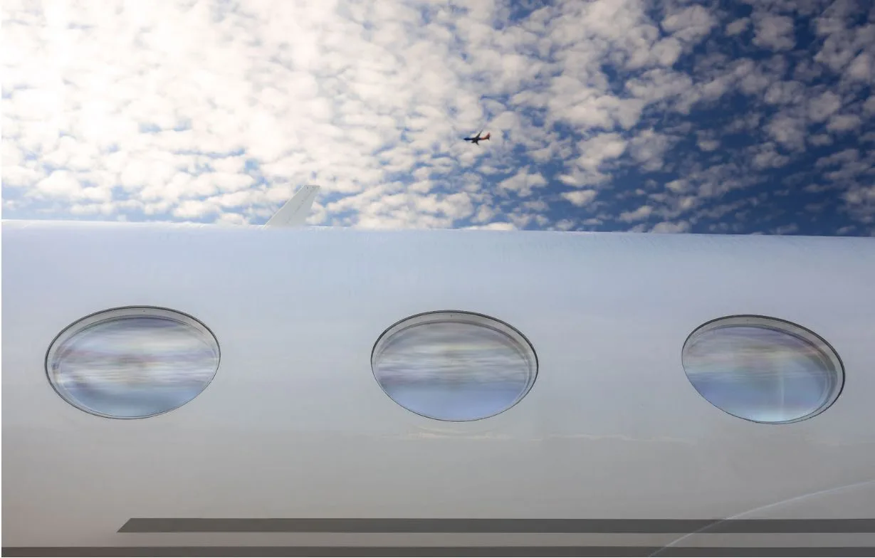 A close-up of three round windows on the white fuselage of a business jet, with another aircraft flying in the distance.