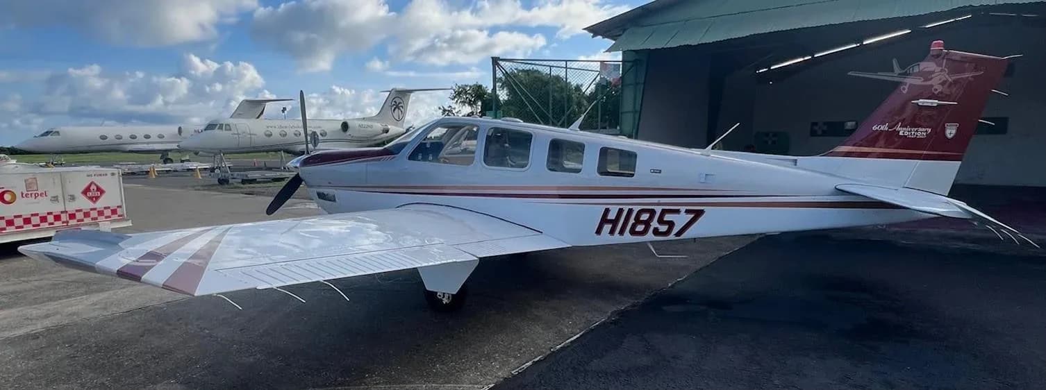 A Beechcraft Bonanza with a silver and maroon paint scheme parked on the tarmac with other aircraft in the background.