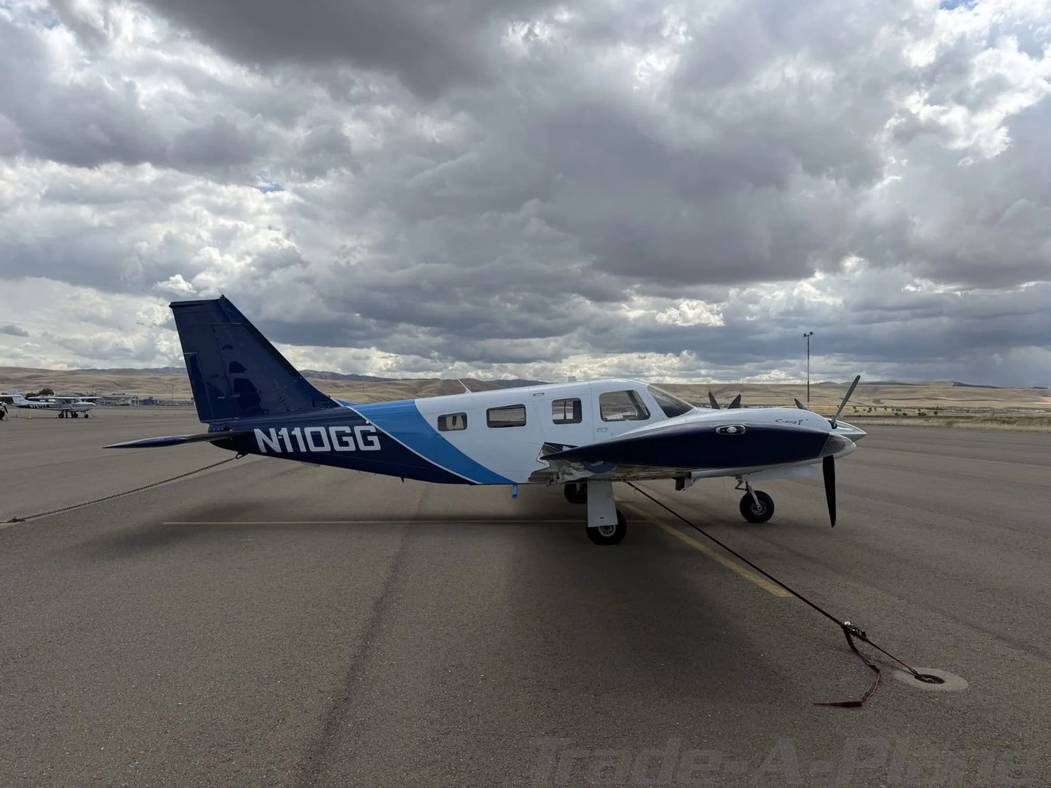 A full side view of the white and blue Piper Seneca II twin-engine aircraft, N110GG, tied down on an airport tarmac under a cloudy sky.