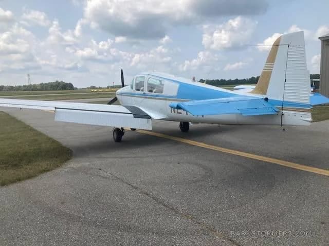 A rear-side view of a white and blue single-engine propeller aircraft with a gold tail, parked on an asphalt ramp.