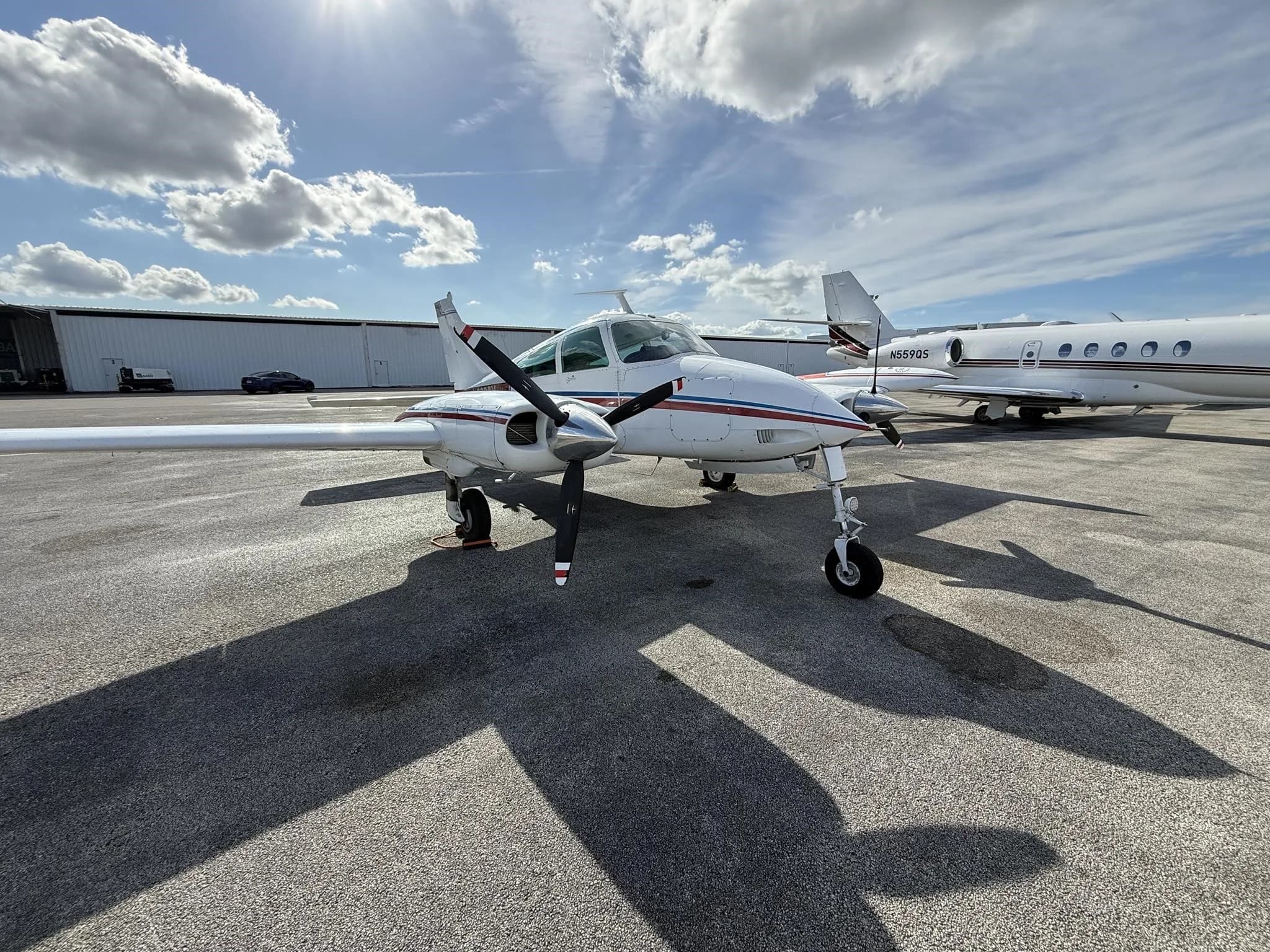 A front three-quarter view of a white Cessna 310L with red and blue stripes parked on an airport tarmac.