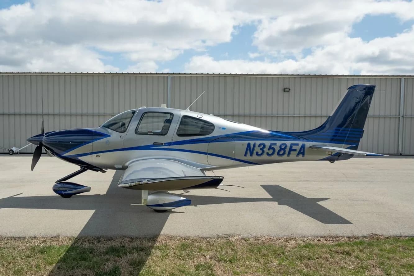 A silver and blue Cirrus SR22T aircraft, registration N358FA, parked on an airfield next to a hangar.