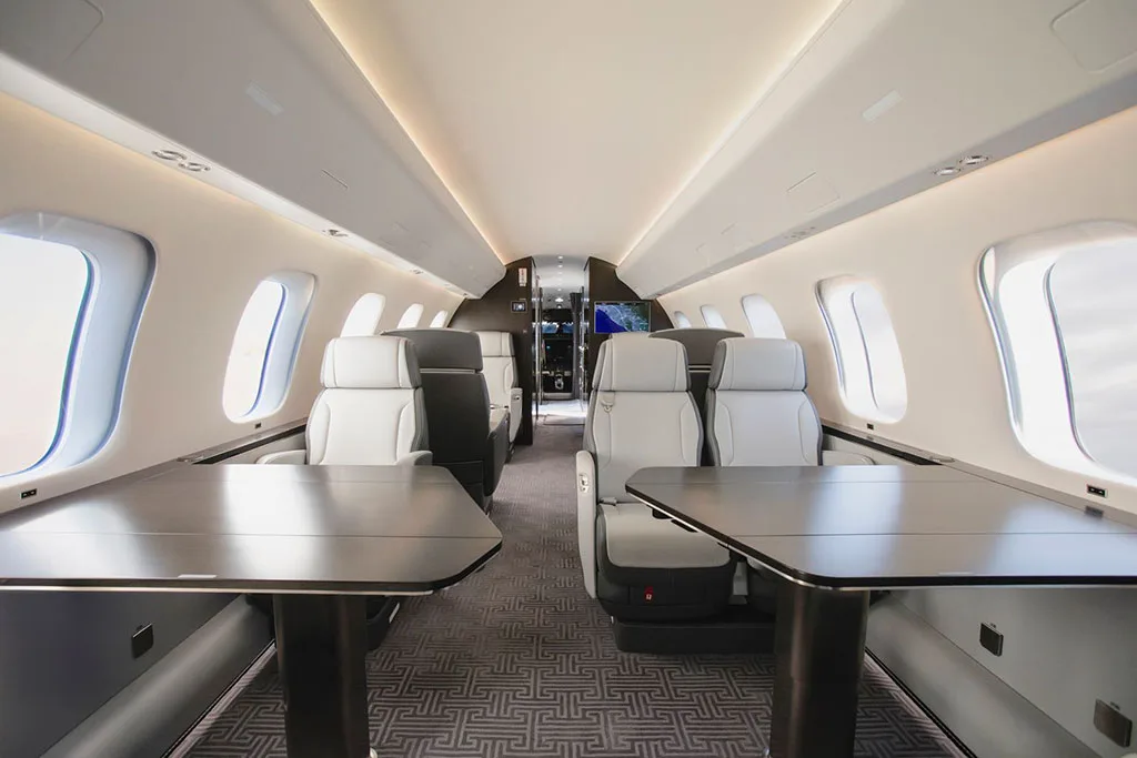 A view down the aisle of a private jet's cabin, featuring light grey and black leather seats with dark tables.