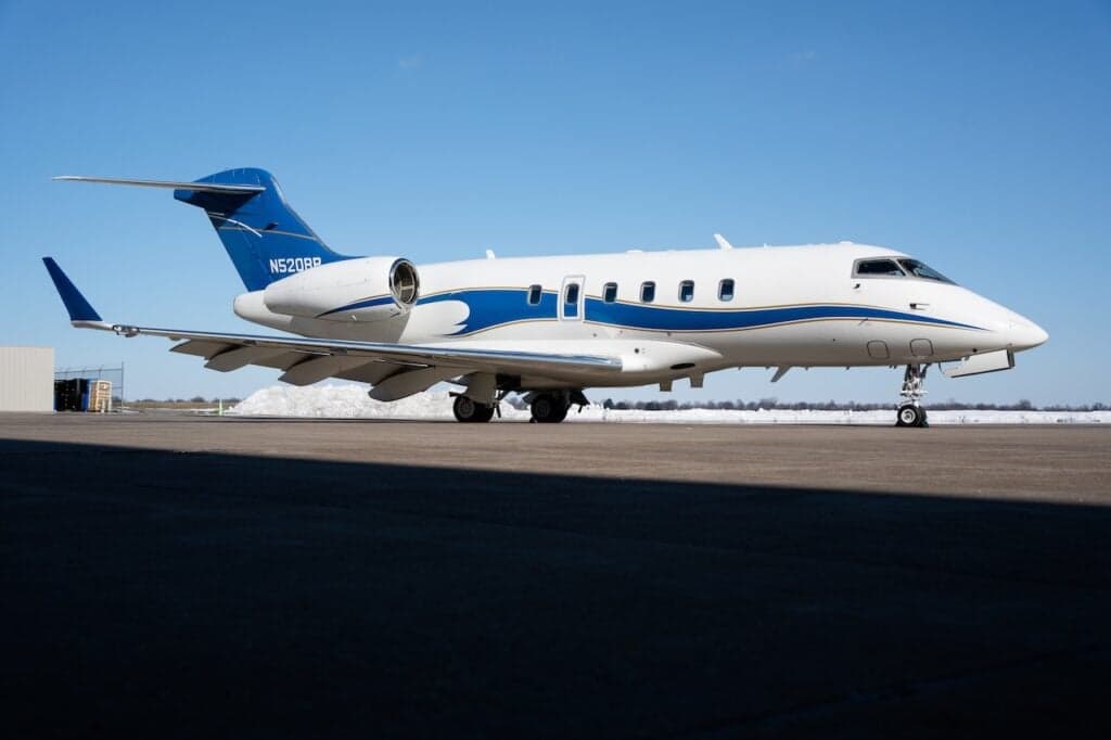 A white and blue Bombardier Challenger 350 business jet with registration N520RP is parked on an airfield under a clear sky.