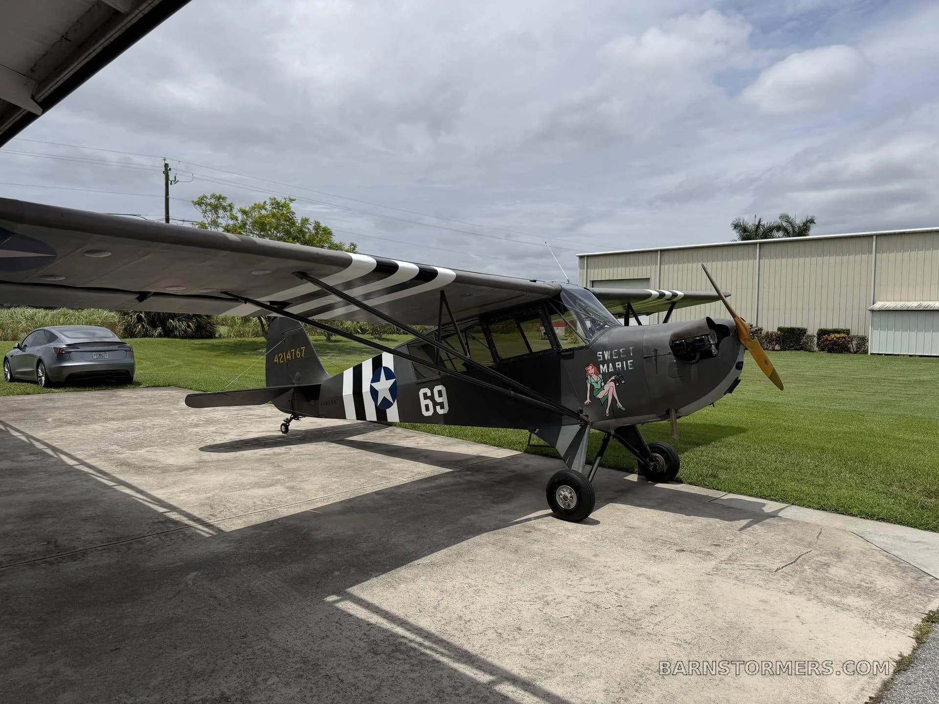 A dark green Piper Cub-style aircraft with "Sweet Marie" nose art and military markings, parked on concrete under a cloudy sky.