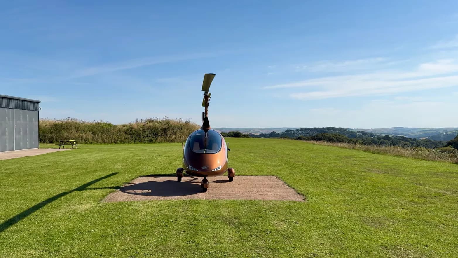 Front view of a brown gyrocopter parked on a concrete pad in a grassy field.