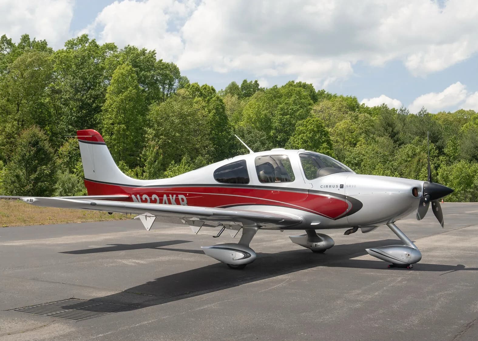 A silver and red Cirrus SR22 aircraft, N224KR, parked on an asphalt ramp with green trees under a partly cloudy sky.