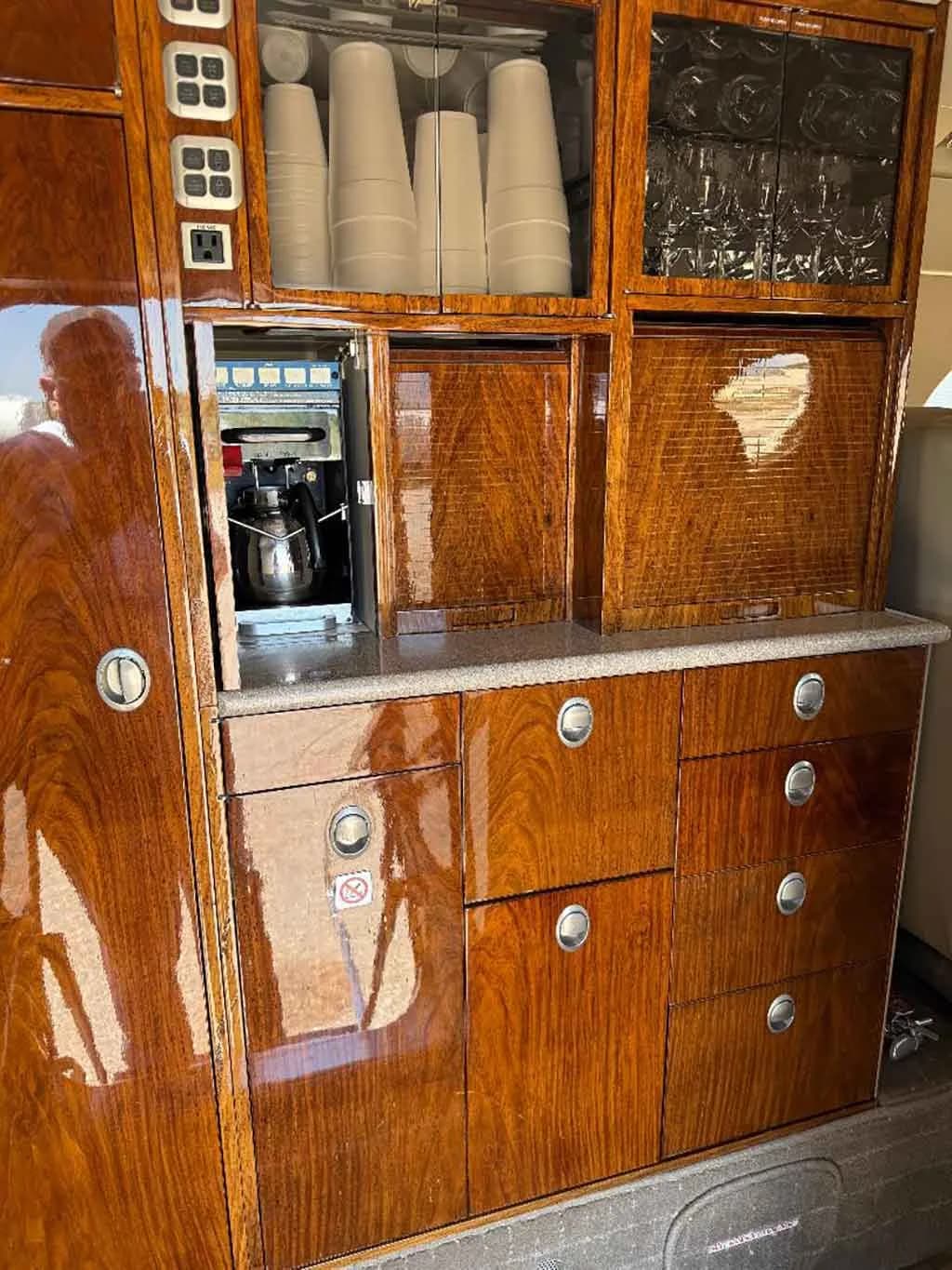 A detailed view of the aircraft's galley, featuring polished wood cabinets, a coffee maker, and storage for cups and glasses.