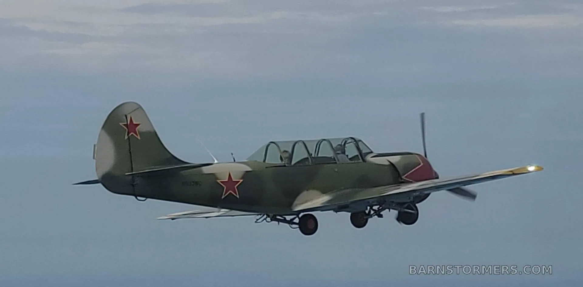 A Yakovlev Yak-52 aircraft in green camouflage with red stars and a red nose, flying against a cloudy sky.