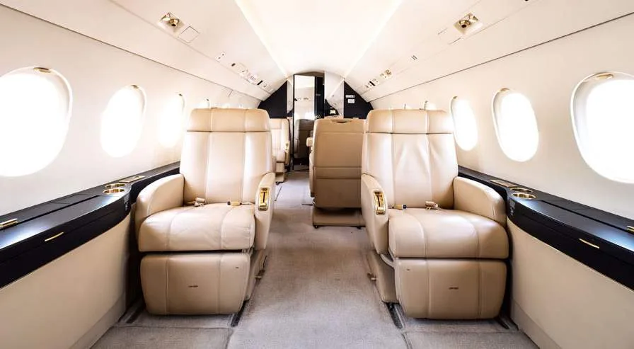 A view down the aisle of a business jet cabin, showing two rows of plush beige leather seats.