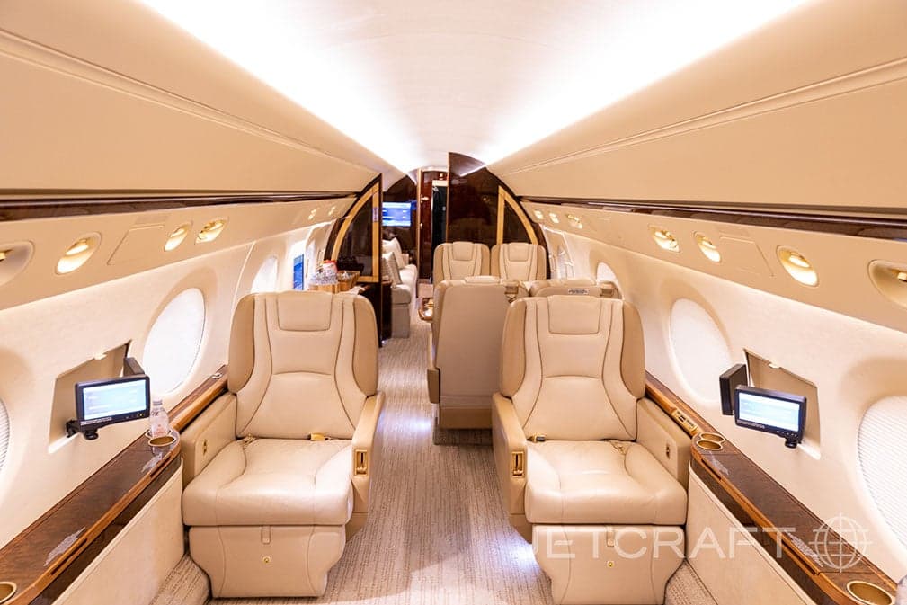The luxurious cabin interior of a Gulfstream G550 business jet with beige leather seats and wood accents.
