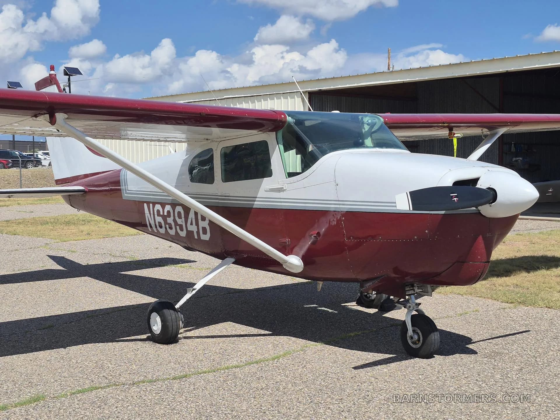 A red and white single-engine propeller aircraft (N6394B) parked on an asphalt ramp next to a hangar under a partly cloudy sky.