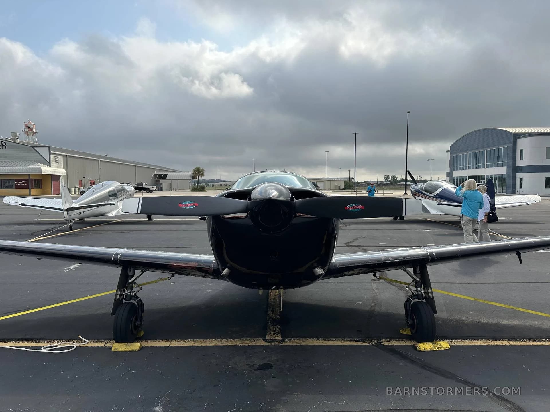 A front view of a black single-engine aircraft with a propeller, parked on an asphalt apron with other aircraft and buildings in the background.