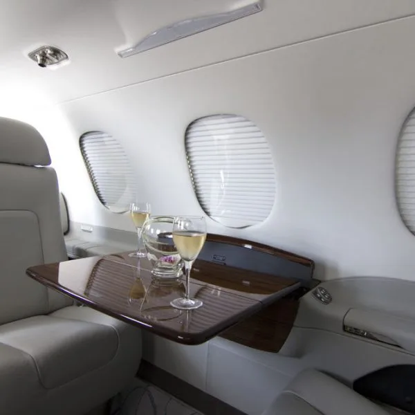 A wooden fold-out table with champagne glasses next to two windows with blinds in the private jet's cabin.