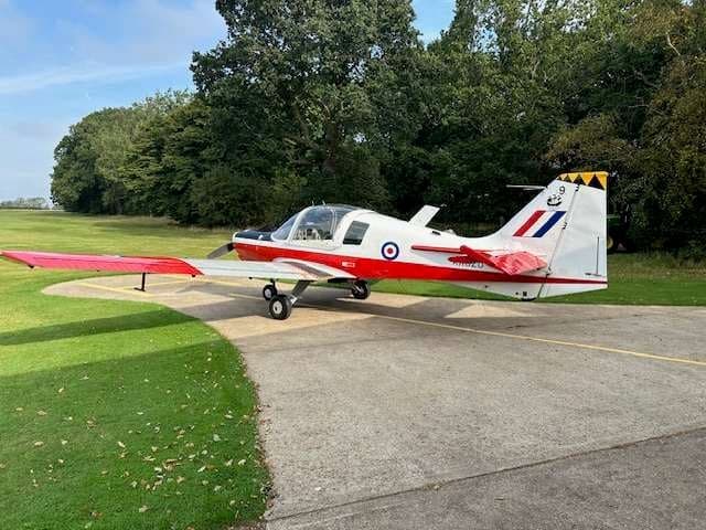 A red and white Bulldog aircraft parked on a paved area next to a grassy field.