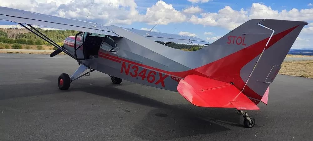 A rear-side view of the gray and red N346X aircraft with "STOL" on its tail, parked on an asphalt apron.