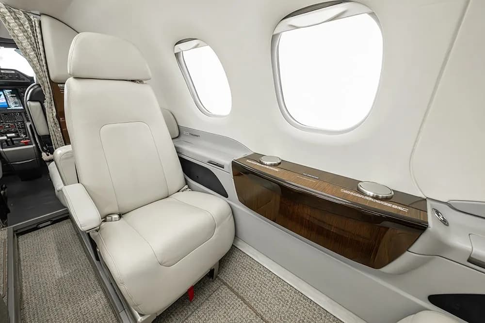 A close-up of a cream leather passenger seat and a polished wooden side table with cup holders in the Embraer Phenom 100E cabin.