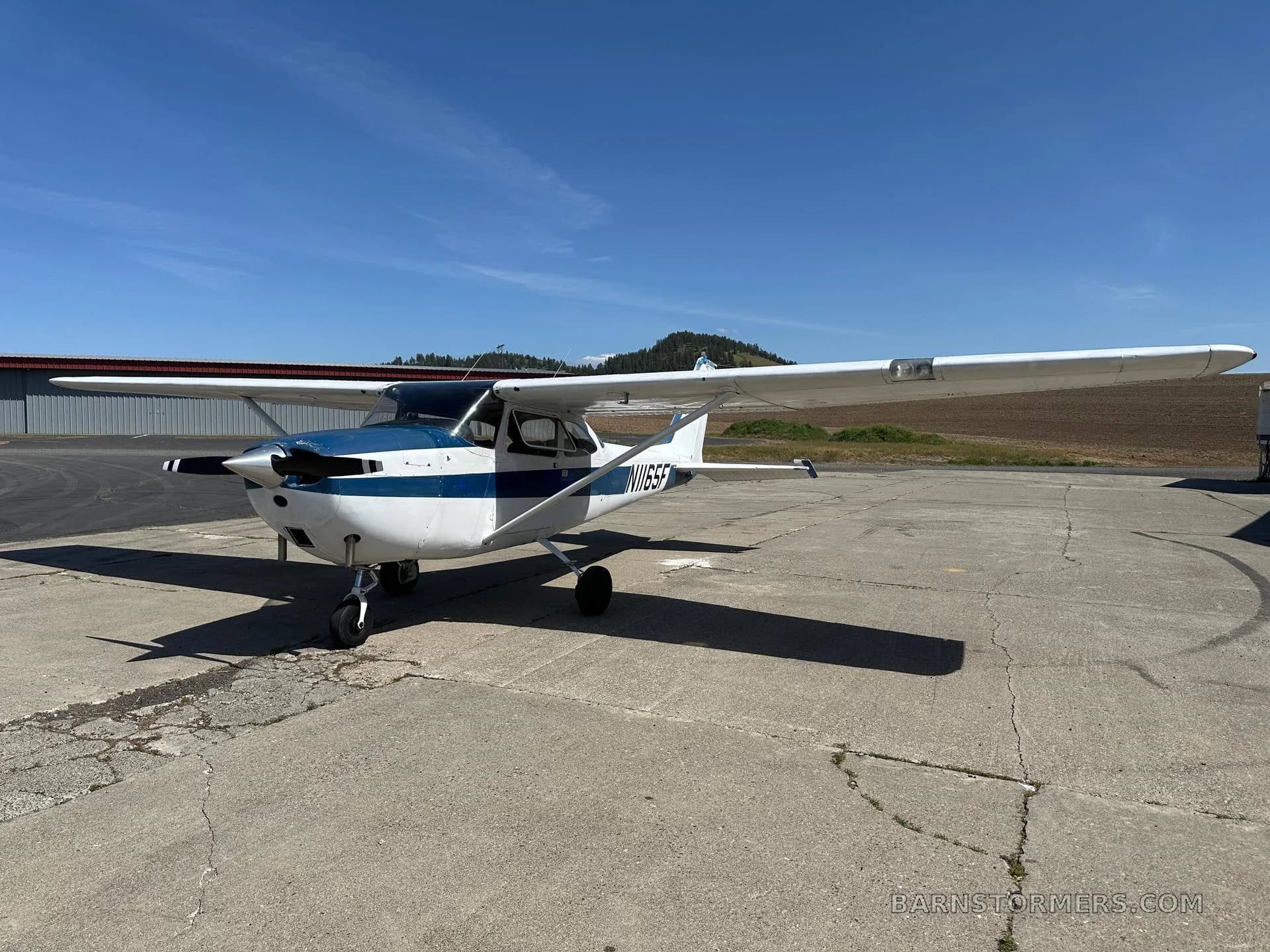 A white and blue Cessna N1165F aircraft is parked on a concrete apron with a field and hills in the background.