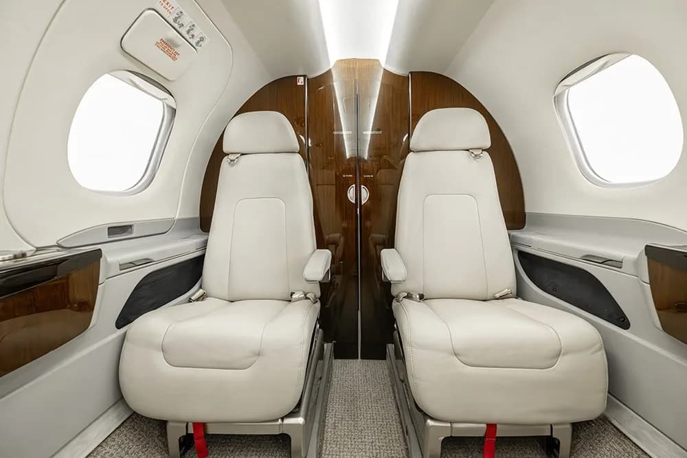 The luxurious interior of the Embraer Phenom 100E cabin, showing two cream leather seats facing a wooden partition.