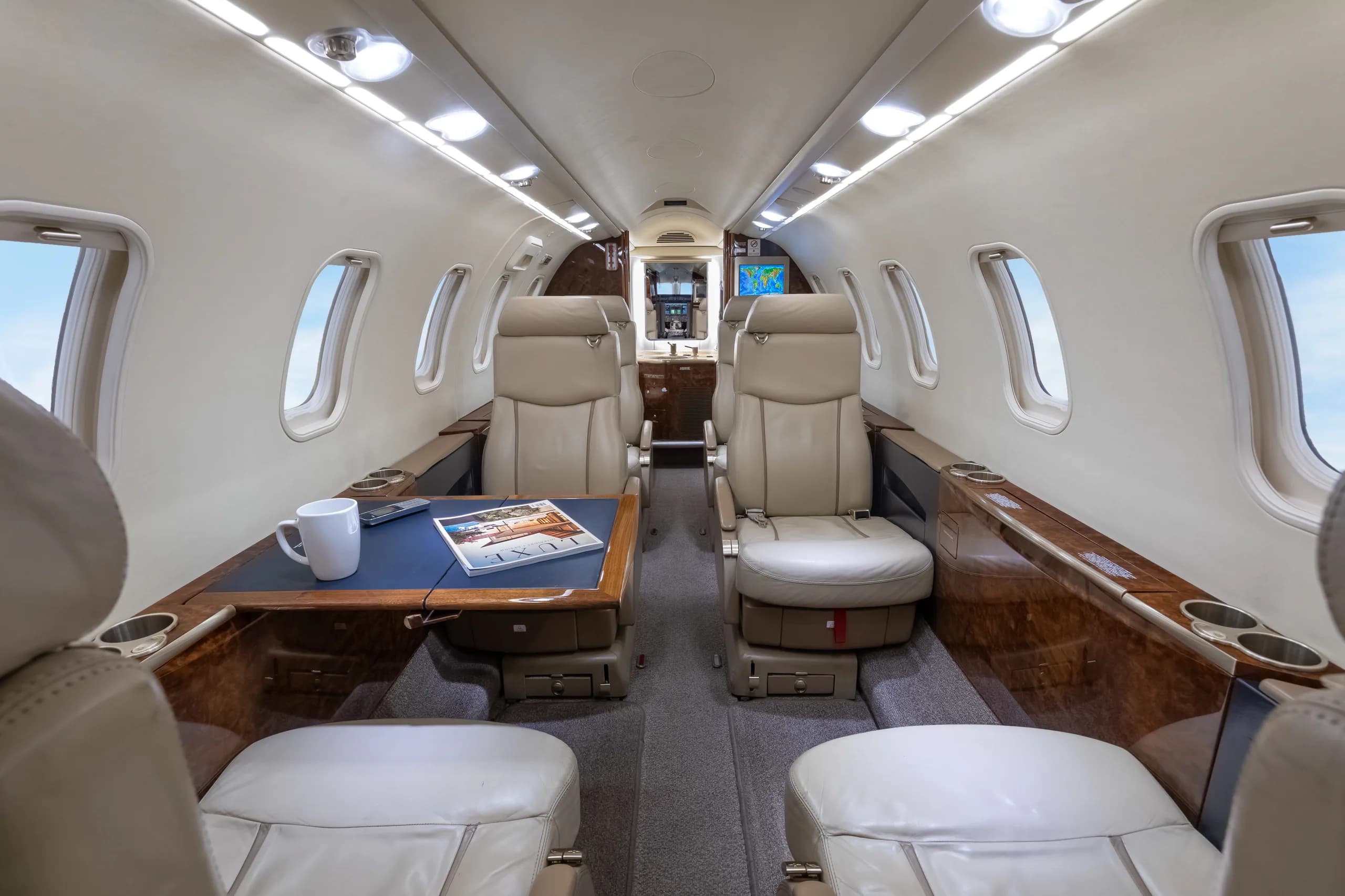 The luxurious cabin interior of a private jet, featuring beige leather seats, a dark wood table, and overhead lighting.