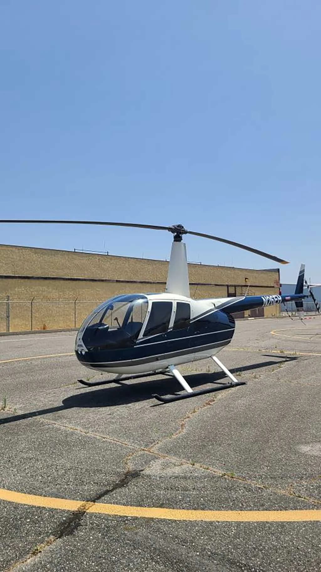 A blue and white Robinson R44 helicopter is parked on an asphalt tarmac with a building in the background.