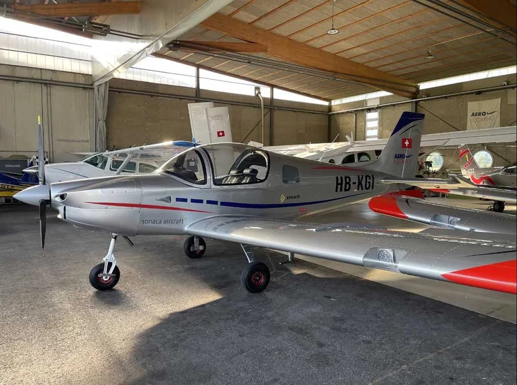 A silver Sonaca 200 aircraft with blue and red stripes, registration HB-KGI, parked inside a hangar, viewed from the front-right.