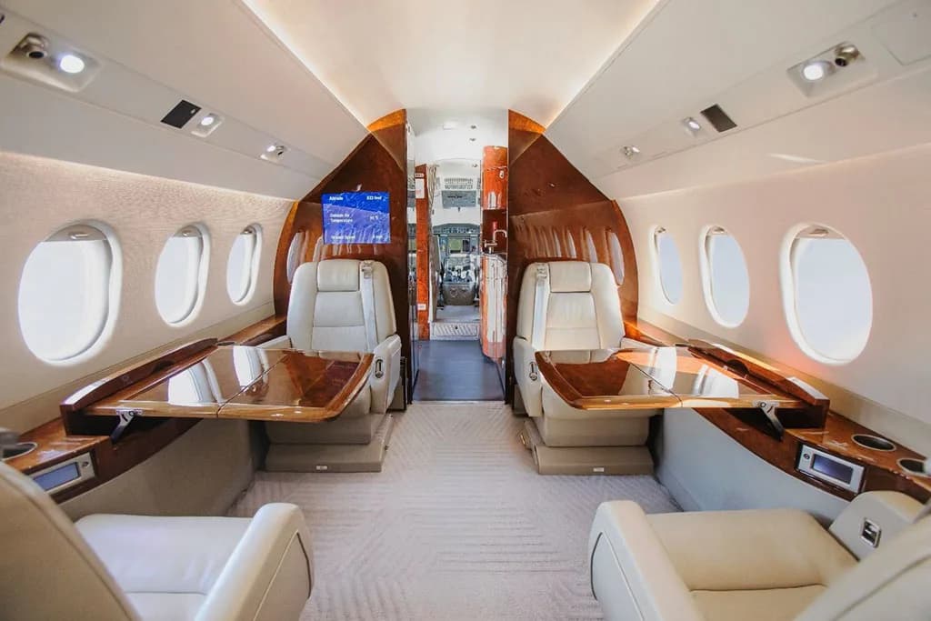 The luxurious interior cabin of a private jet, featuring light leather seats, polished wood tables, and a screen displaying flight information.