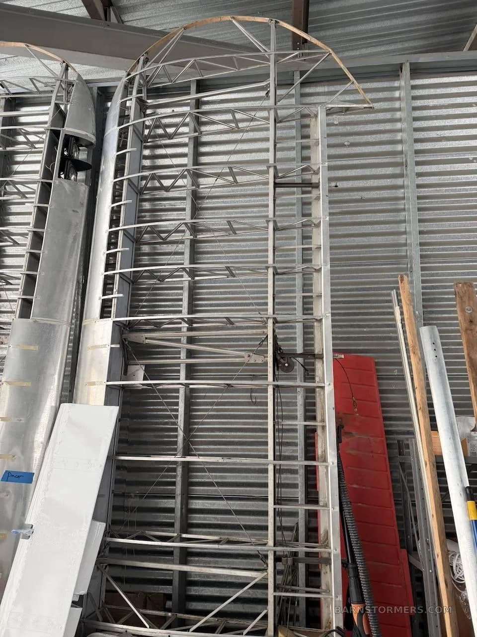 Aircraft wing and fuselage frame components, including a red control surface, stored in a metal hangar.