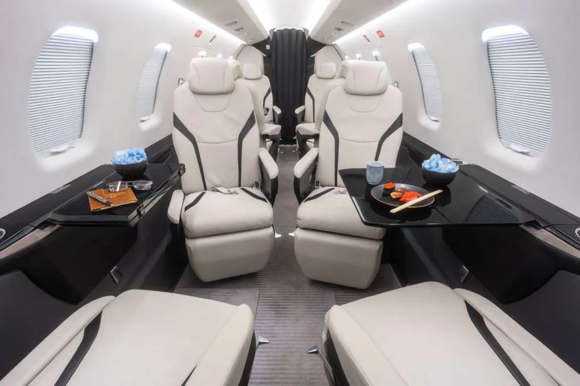 The elegant interior cabin of a private jet shows white leather seats, fold-out tables with snacks, and large windows.