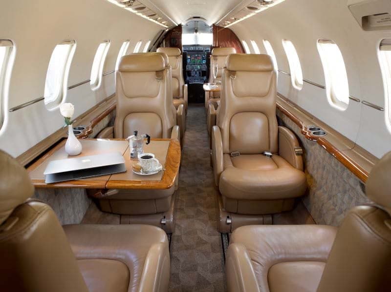 A full view of the aircraft cabin interior, featuring tan leather seats, wooden tables, and a laptop with coffee.