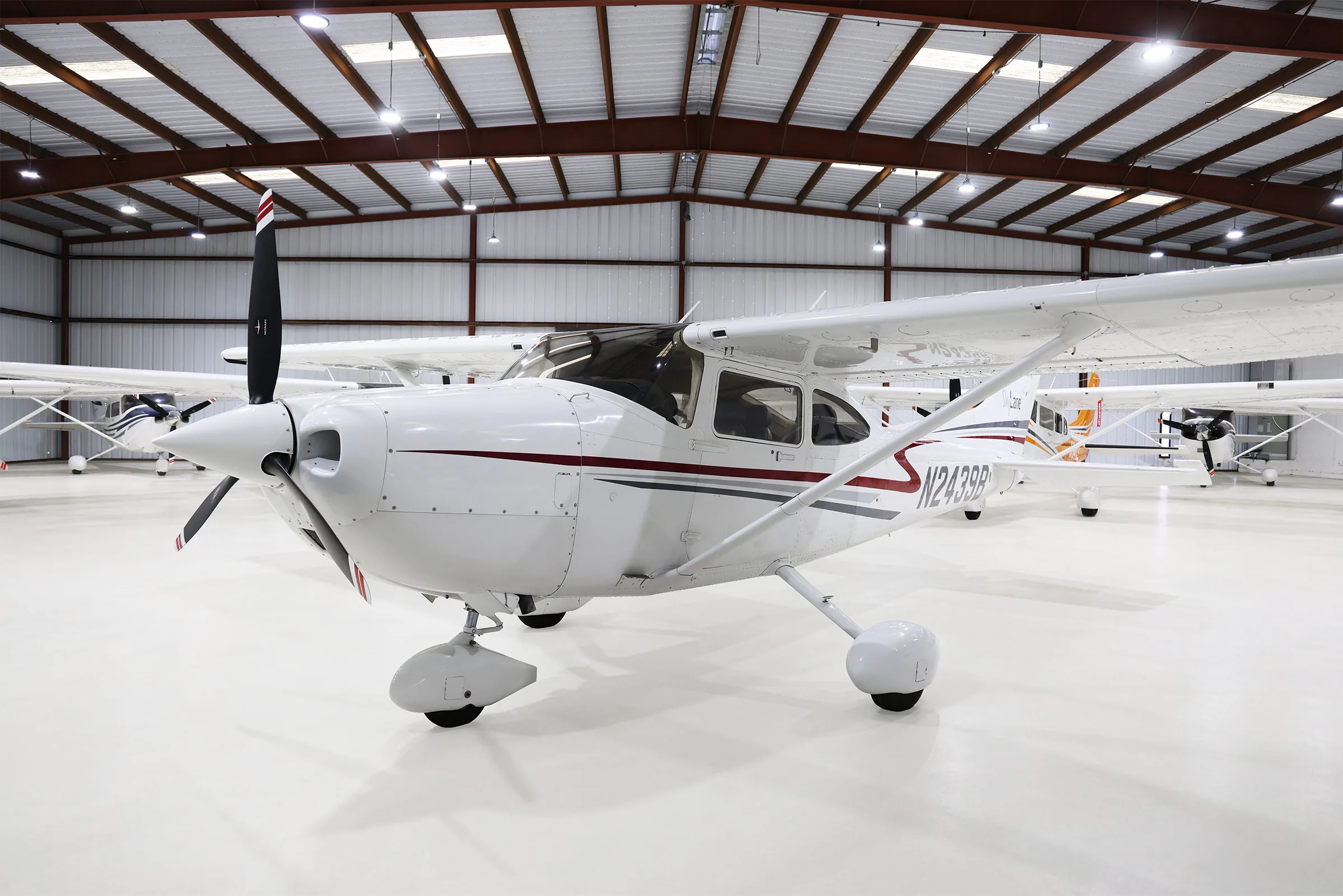 A white Cessna 182 Skylane with red and grey stripes, parked in a hangar, viewed from the front-left.