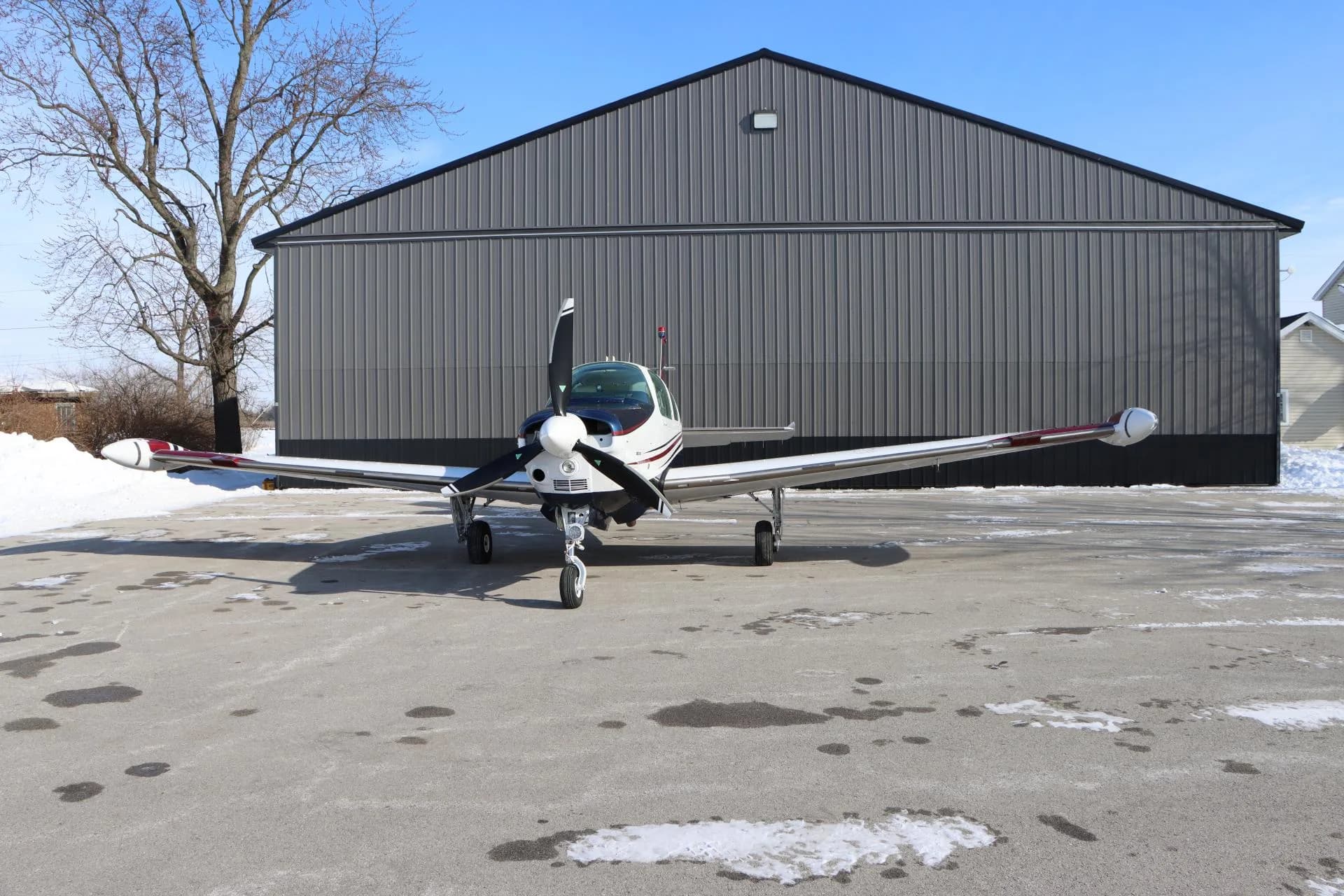 Front view of a white, blue, and red single-engine aircraft parked on a partially snowy tarmac in front of a hangar and bare trees.