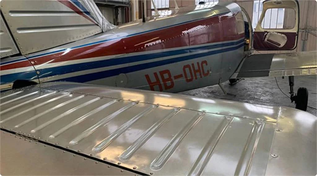 View of the wing and rear fuselage of a red and silver aircraft with blue stripes and registration HB-OHC, with an open door.