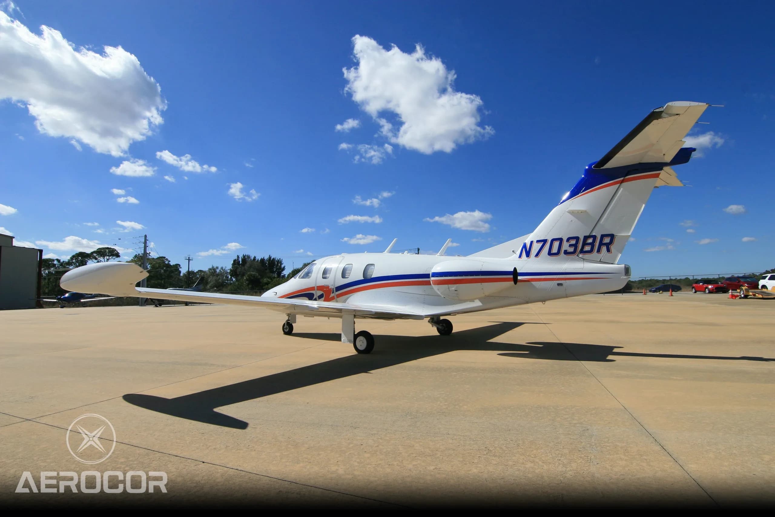 A side view of a white Eclipse 500 jet with blue and orange stripes, tail number N703BR, parked on an airfield under a partly cloudy sky.